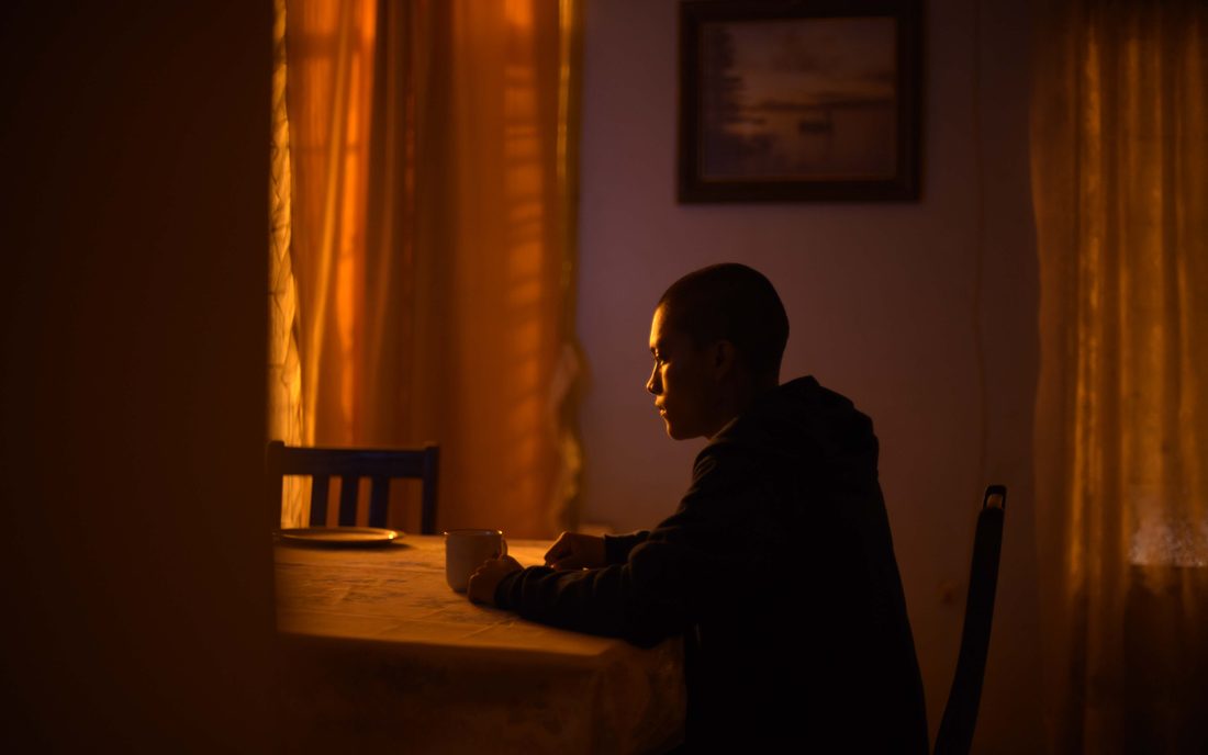 Fotograma de Vergüenza: un joven sentado a la mesa en una cocina a oscuras iluminada por una lámpara ámbar